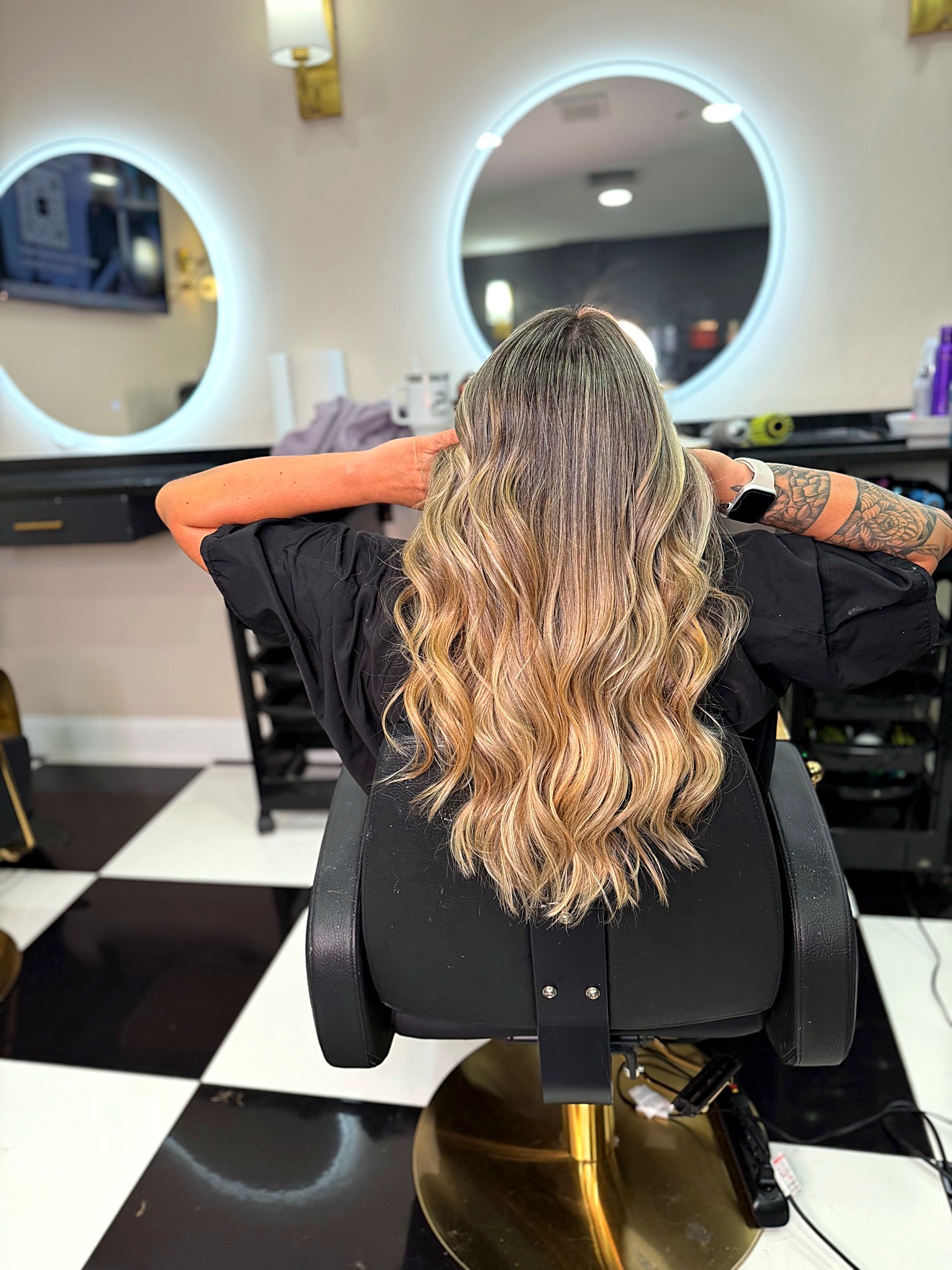 Blonde long wavy hair, back view, seated in a salon chair with round mirrors